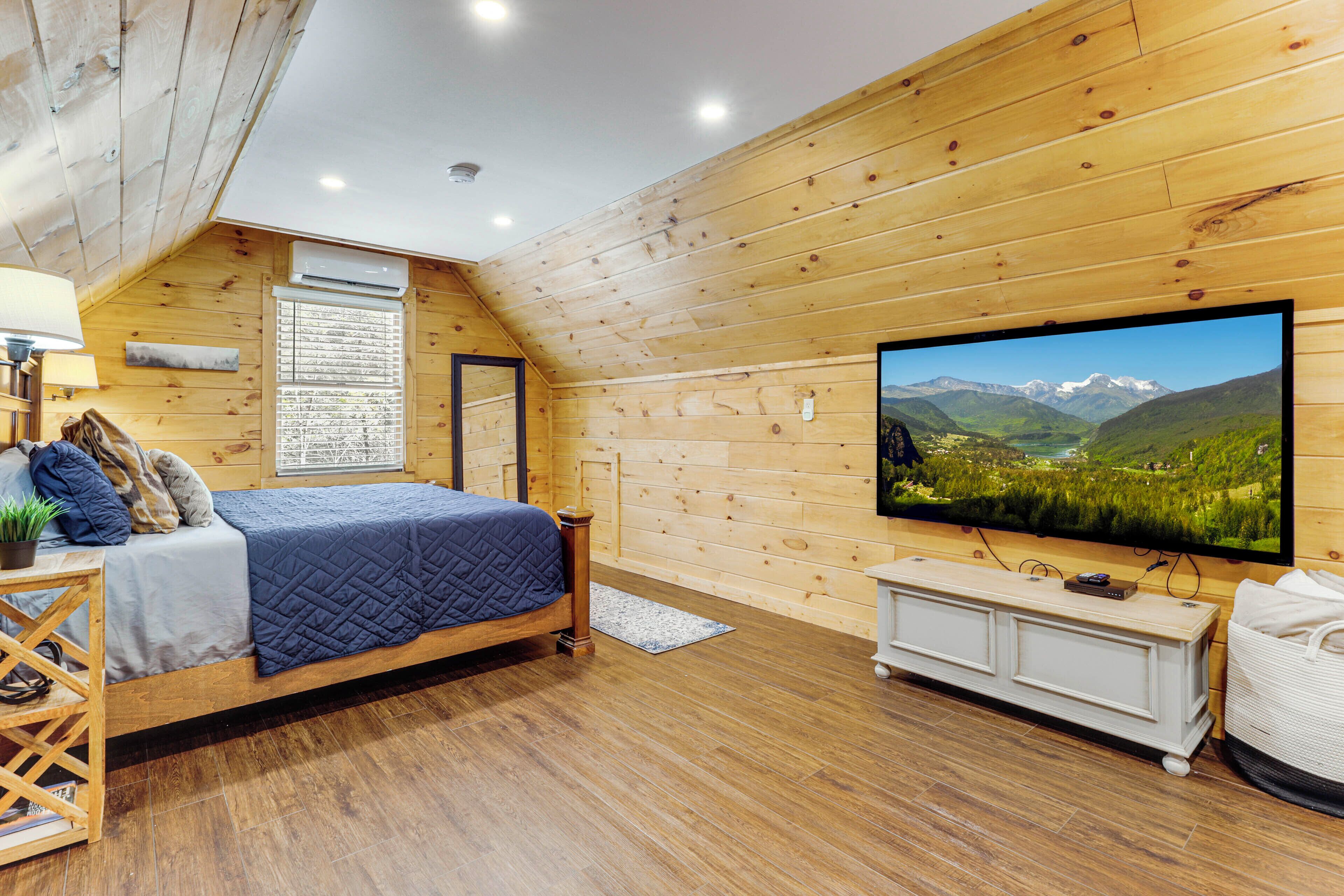 A cozy bedroom with a wooden sloped ceiling, a large bed with a blue quilt, a mounted TV displaying a scenic landscape, and a white dresser.