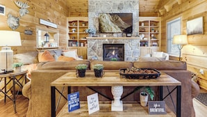 A cozy log cabin interior with a stone fireplace, a television mounted above it, and a comfortable seating area with a sofa and a coffee table. - Waterfall Log Cabin: Private Hot Tub & Game Room (Gatlinburg)