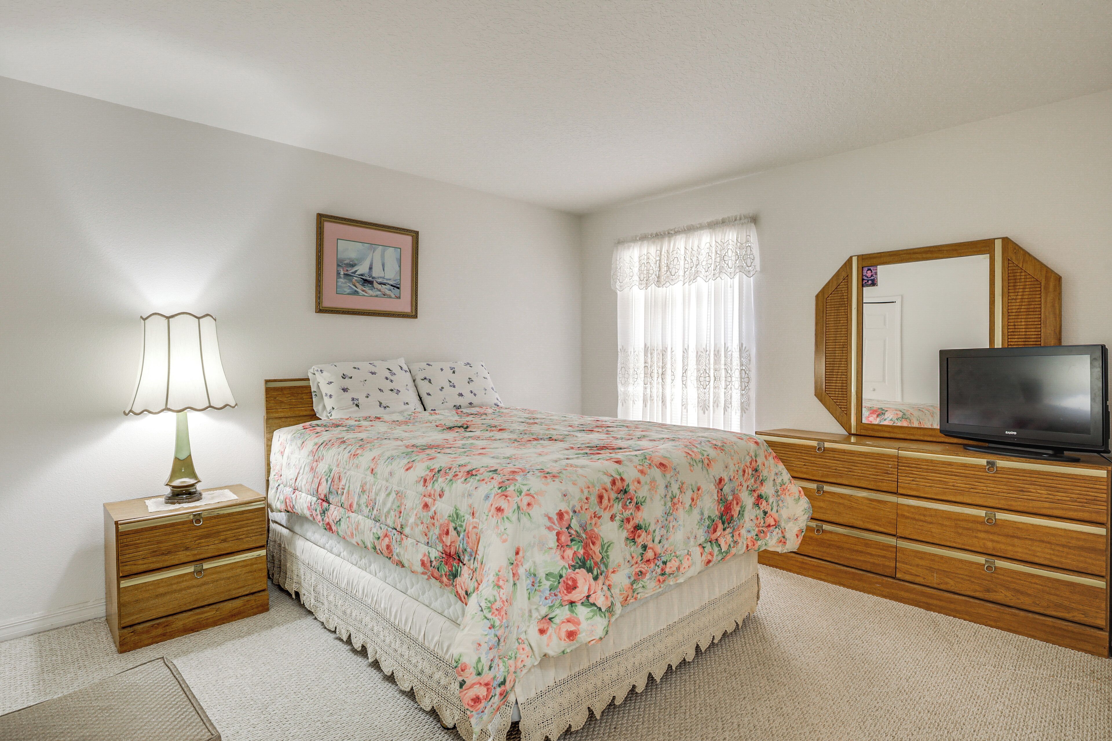 A bedroom with a bed, a nightstand with a lamp, a dresser, a television, and a window with curtains.