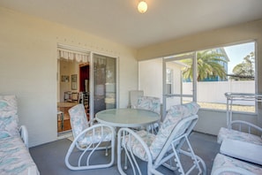 Un patio cubierto con una mesa redonda de vidrio y sillas de mimbre blanco, una puerta corrediza de vidrio que da a un espacio interior, y vista a un edificio azul y palmeras afuera.