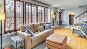 A spacious living room with a beige sofa, ottoman, and a small metal shelf. Large windows with wooden frames provide ample natural light. - Lakeside Clifton Heights Home Near Forest Park (St. Louis)