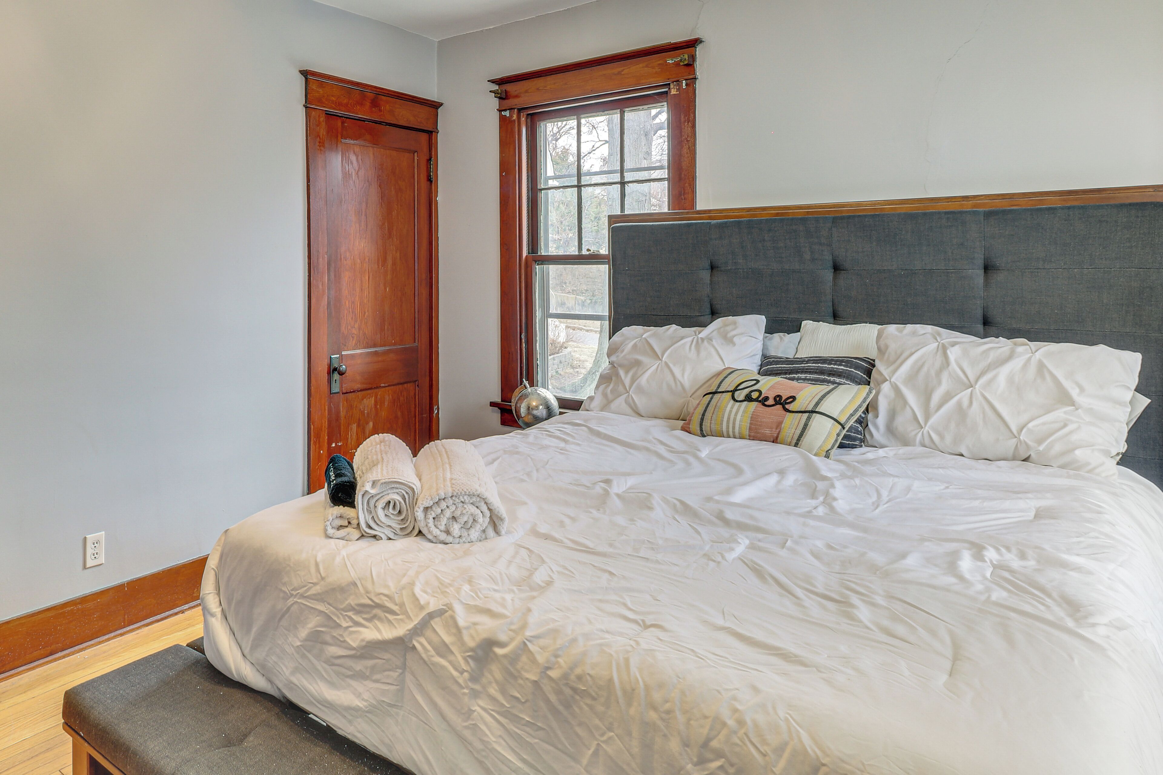 A neatly made bed with white linens, a bench at the foot of the bed, a wooden door, and a window with a view of trees.