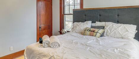 A neatly made bed with white linens, a bench at the foot of the bed, a wooden door, and a window with a view of trees.