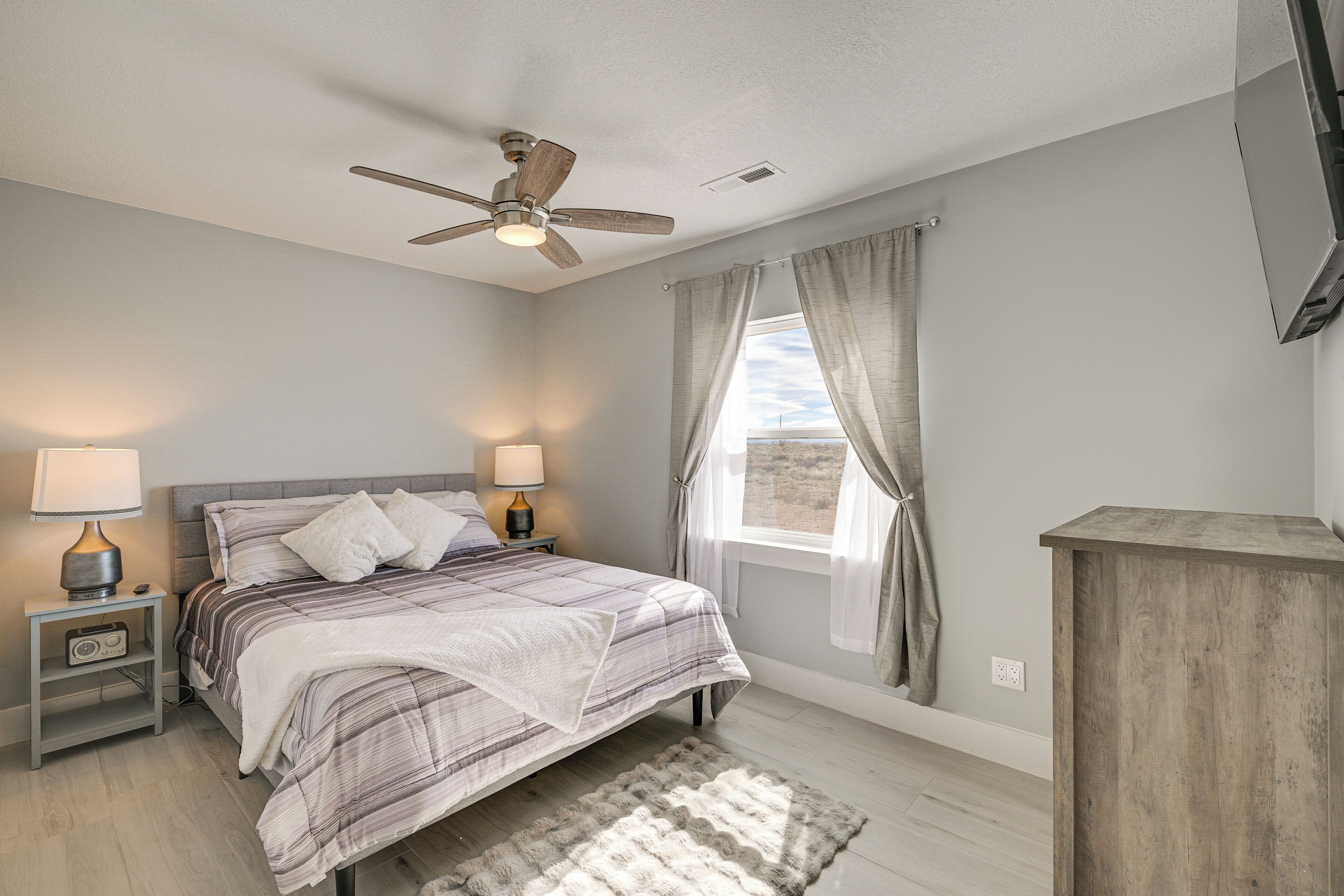 Une chambre à coucher comprenant un lit, des tables de chevet, un ventilateur de plafond, une fenêtre avec des rideaux et une commode en bois.