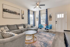 A living room with a gray sectional sofa, a round coffee table, and a blue patterned rug. There is a ceiling fan, a door, and a window with blue curtains. - 1 Mi to Downtown Greensboro: Newly Built Home! (Greensboro)