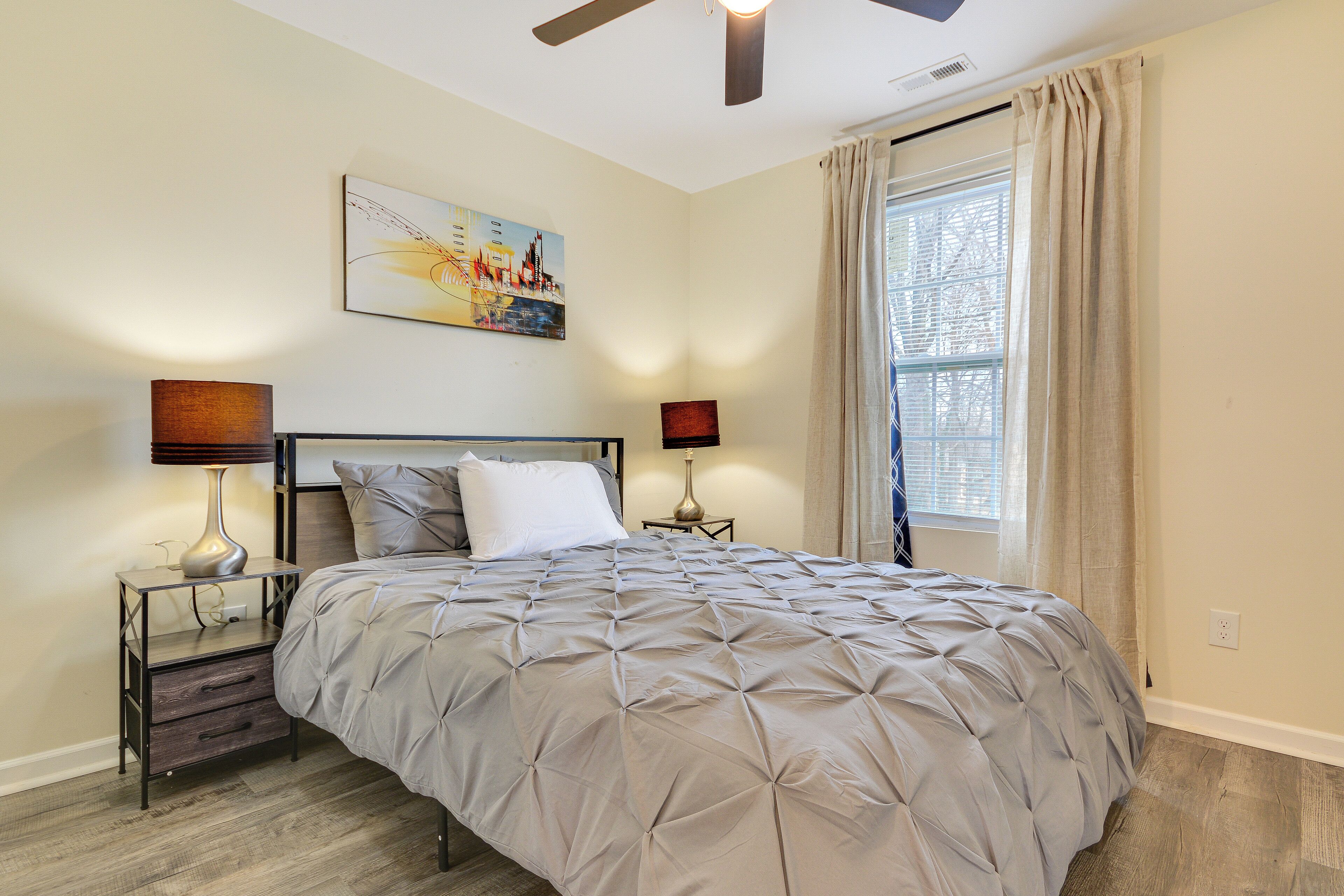A bedroom with a bed, two bedside tables, a ceiling fan, a window with curtains, and a painting on the wall.