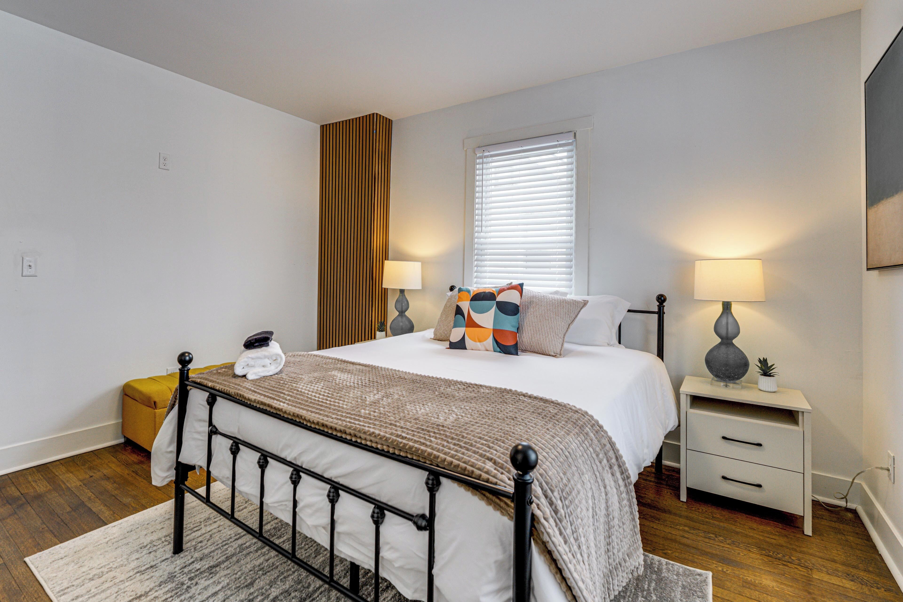 Une chambre à coucher comprenant un lit, des tables de chevet, une commode, une fenêtre avec des stores et un mur d’accent en bois.
