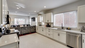 A modern kitchen with white cabinets, a stainless steel dishwasher, and a gray sofa in the living area. - Spacious & Updated Group Getaway in Detroit! (Detroit)