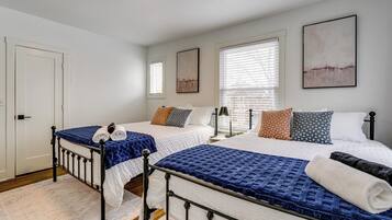 Une chambre à coucher avec deux lits, chacun recouvert d’une couverture bleue, avec du linge de lit blanc et des oreillers à motifs. On y trouve deux tables de chevet avec des lampes, une porte et une fenêtre avec des stores.