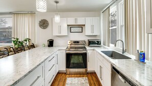 Una cocina moderna con armarios blancos, electrodomésticos de acero inoxidable, un amplio ventanal y una lámpara colgante.