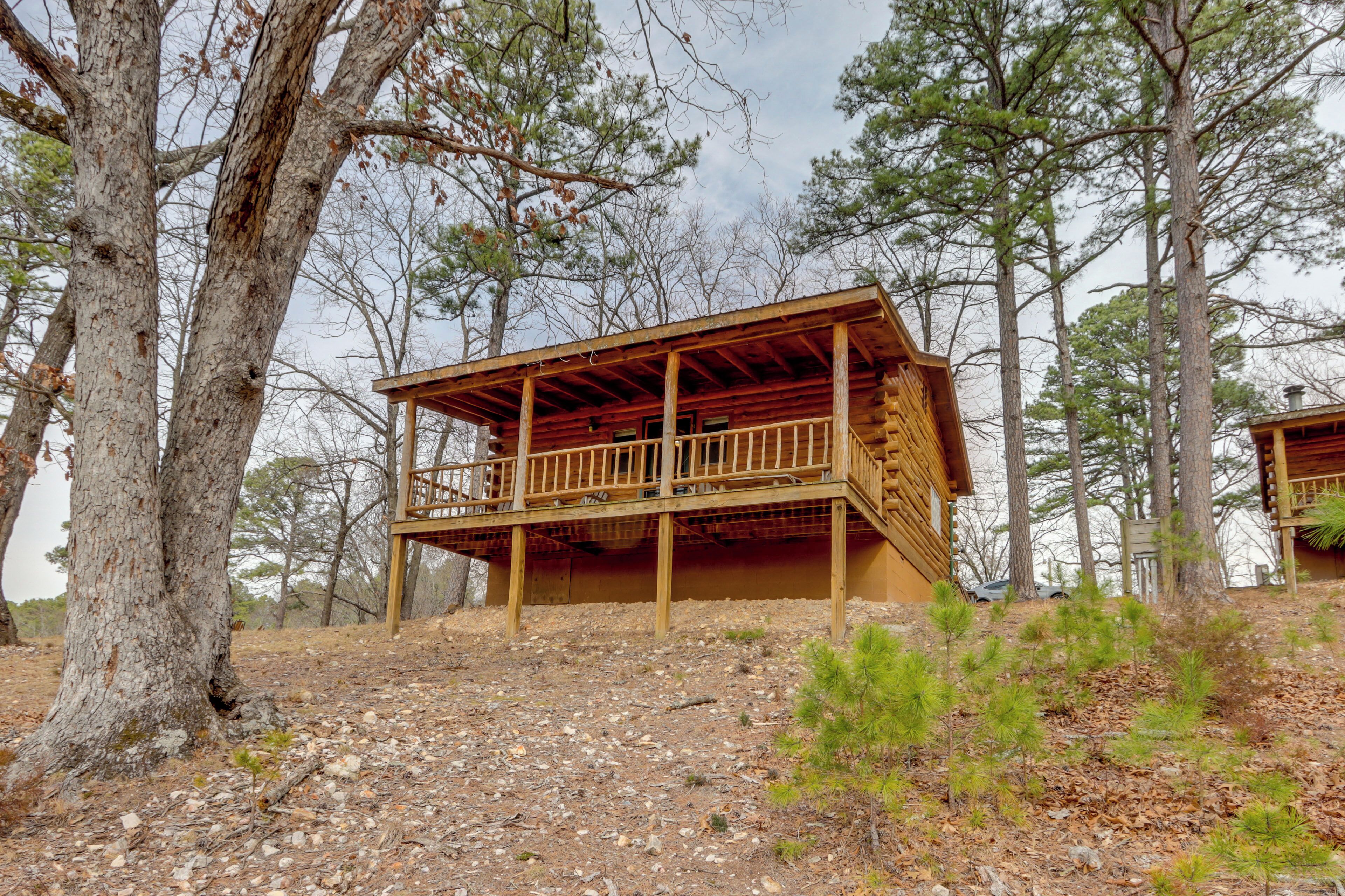 A wooden cabin with a balcony, surrounded by trees and a rocky ground.
