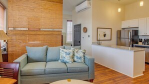 Un salon moderne avec un canapé gris, un mur recouvert de panneaux en bois et un coin cuisine doté de meubles blancs et d’appareils électroménagers en acier inoxydable.