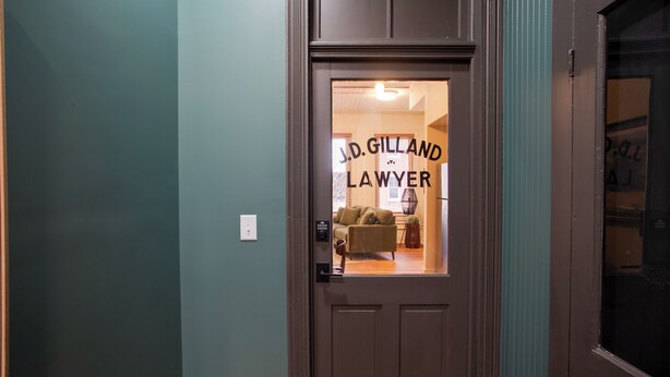 Een deur met een bord waarop staat 'J.D. GILLAND ADVOCAAT' en een blik op een kamer met een bank en een lamp.