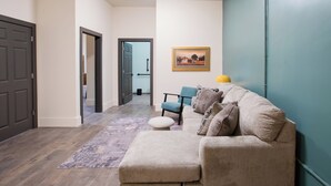 Ein Wohnzimmer mit einem beigen Sofa, einem blauen Sessel und einem kleinen Hocker. An der Wand hängt ein gerahmtes Bild und auf einem Beistelltisch steht eine gelbe Lampe.