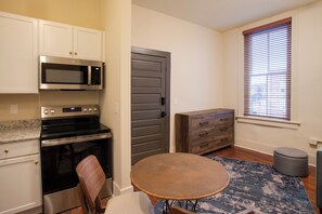 A compact kitchen with a microwave, stove, and cabinets. There's a dining table with a rug and a window with blinds.
