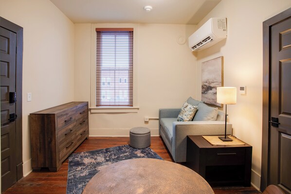 A compact living space with a sofa, a small side table, a lamp, a wooden dresser, and a window with blinds.