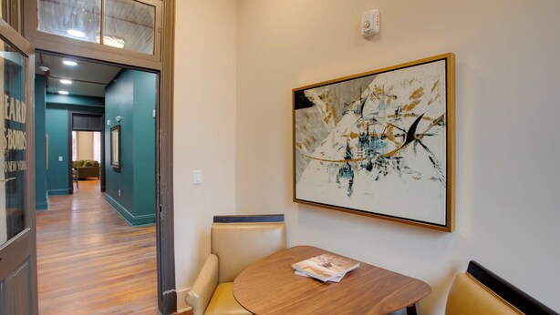 Un hall d’hôtel avec un plancher en bois, une table ronde sur laquelle repose un magazine, et un grand tableau abstrait accroché au mur.
