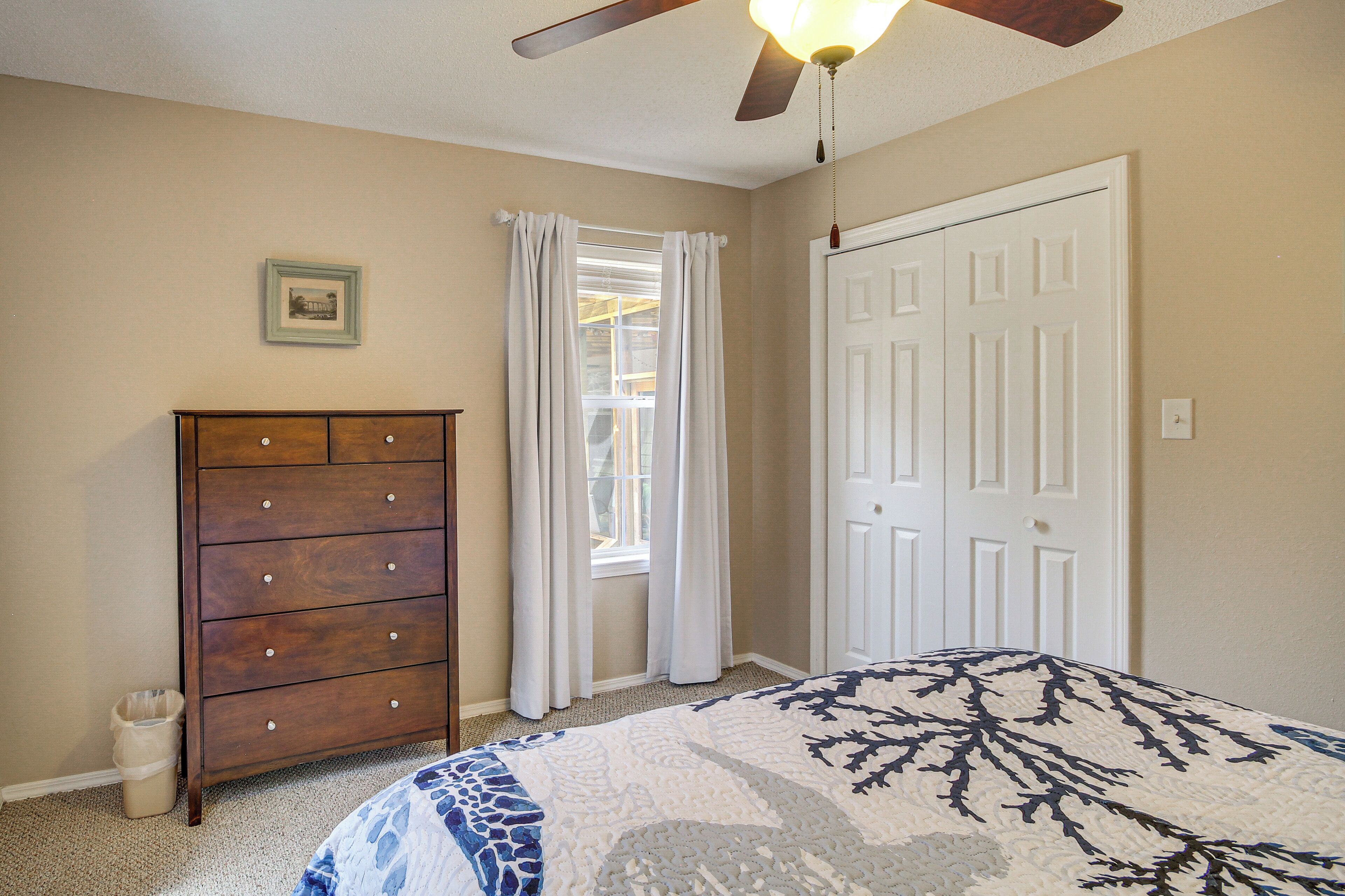 A bedroom with a wooden dresser, a bed with a patterned bedspread, a window with white curtains, and a ceiling fan.