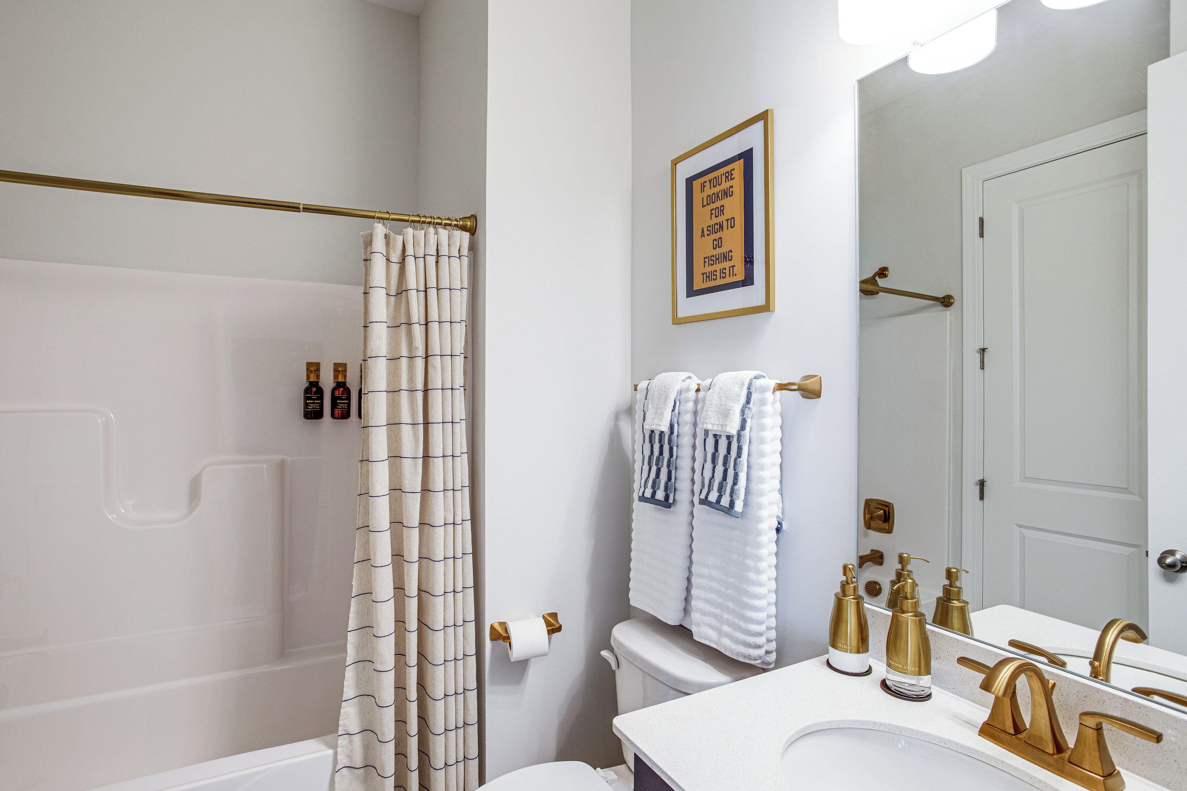A bathroom with a shower curtain, toilet, sink with gold fixtures, and a framed sign on the wall.