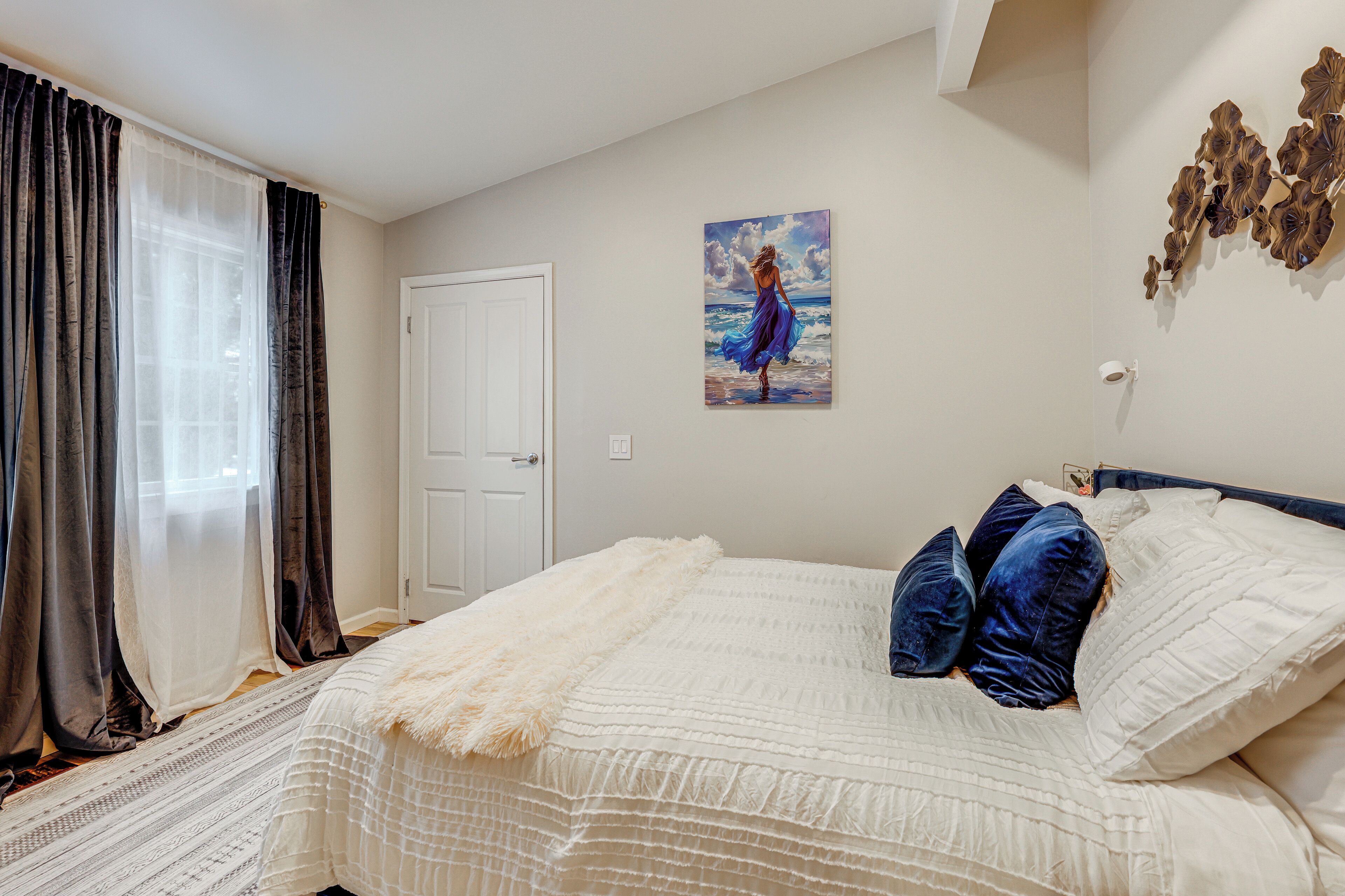 A bedroom with a bed, a white door, a painting, and a wall decoration.