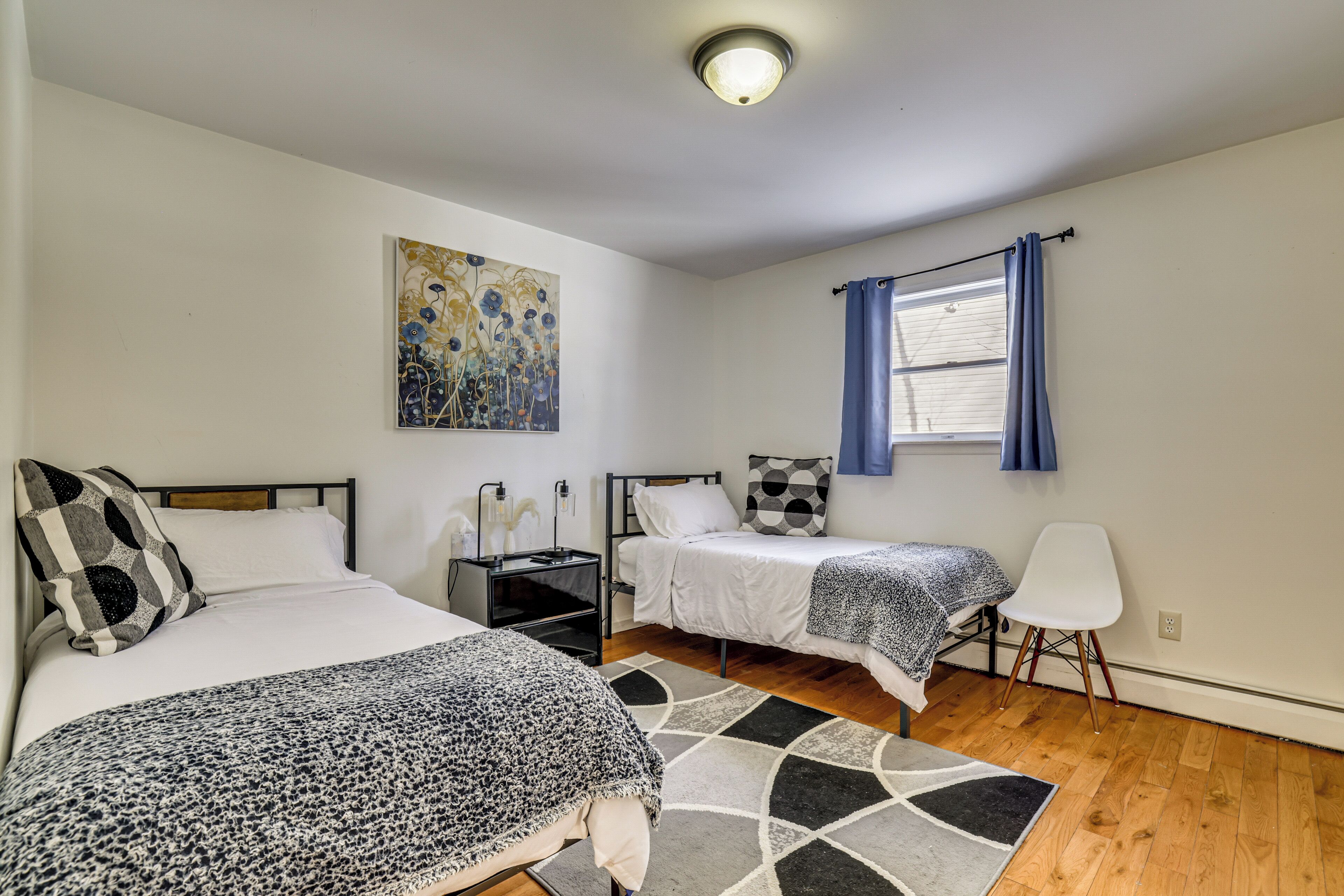 A bedroom with two beds, a nightstand, a chair, a painting, and a window with curtains.