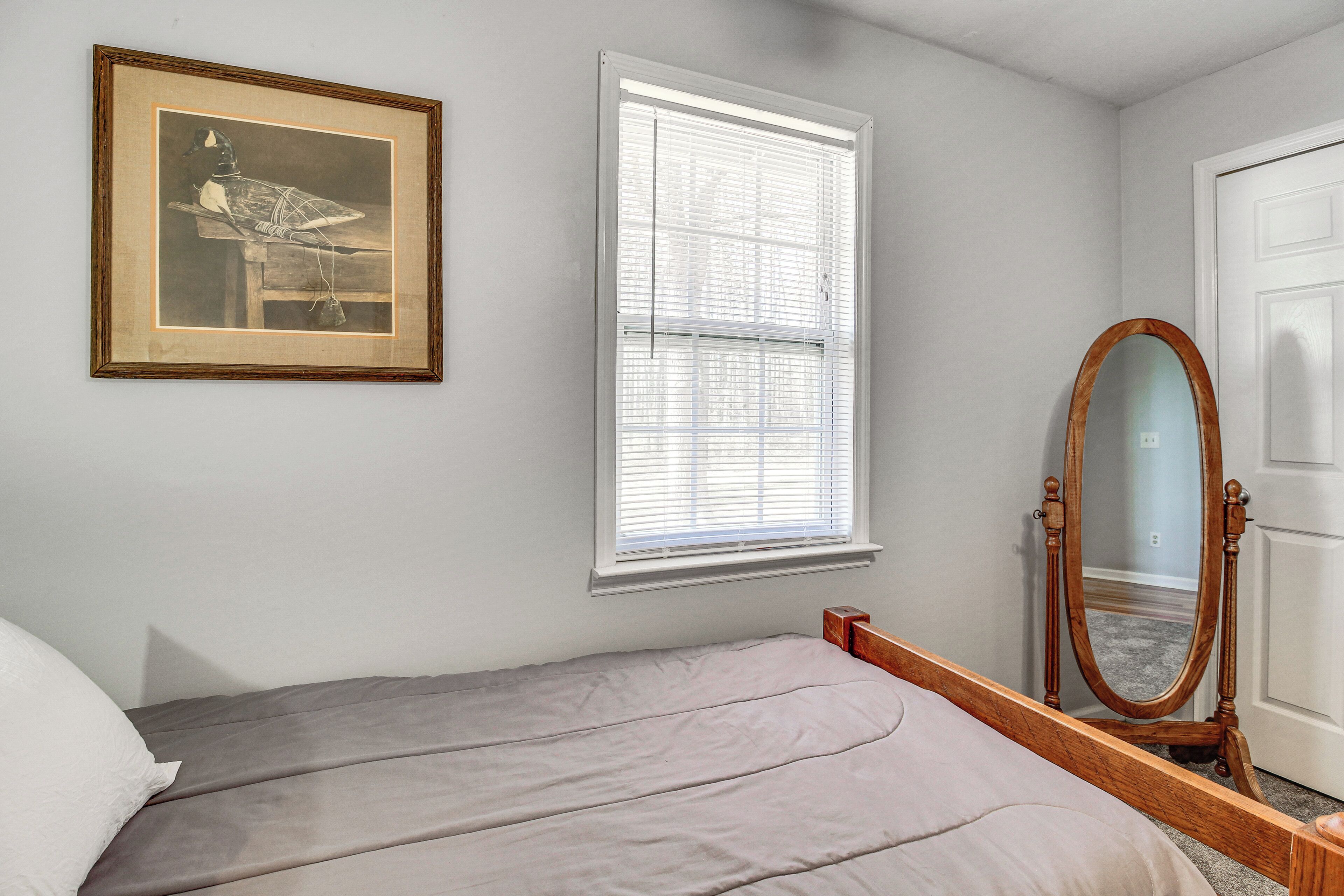 A bedroom with a bed, a framed picture on the wall, a window with blinds, and a wooden-framed mirror.