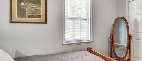 A bedroom with a bed, a framed picture on the wall, a window with blinds, and a wooden-framed mirror.