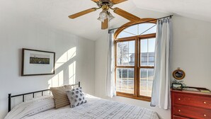 Une chambre à coucher avec un lit, un ventilateur de plafond en bois, une fenêtre avec des rideaux, une commode et un tableau encadré accroché au mur.