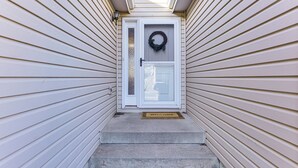Une entrée de maison avec une porte blanche, un paillasson et une guirlande.