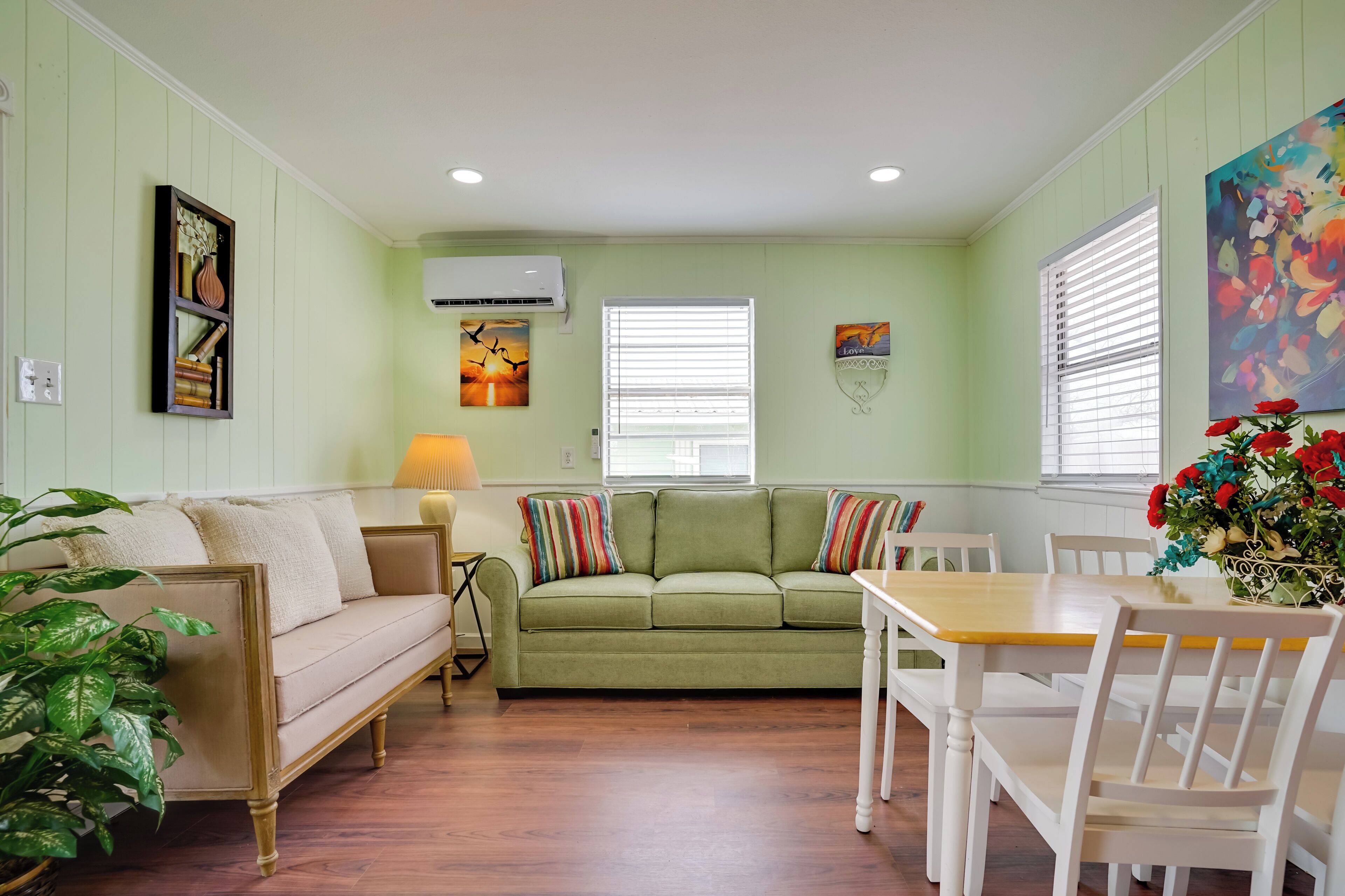 A room with a green sofa, a dining table with chairs, a lamp, and a potted plant.
