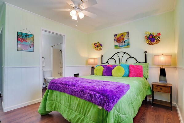A bedroom with a green bedspread, purple and green pillows, a ceiling fan, and two bedside lamps.