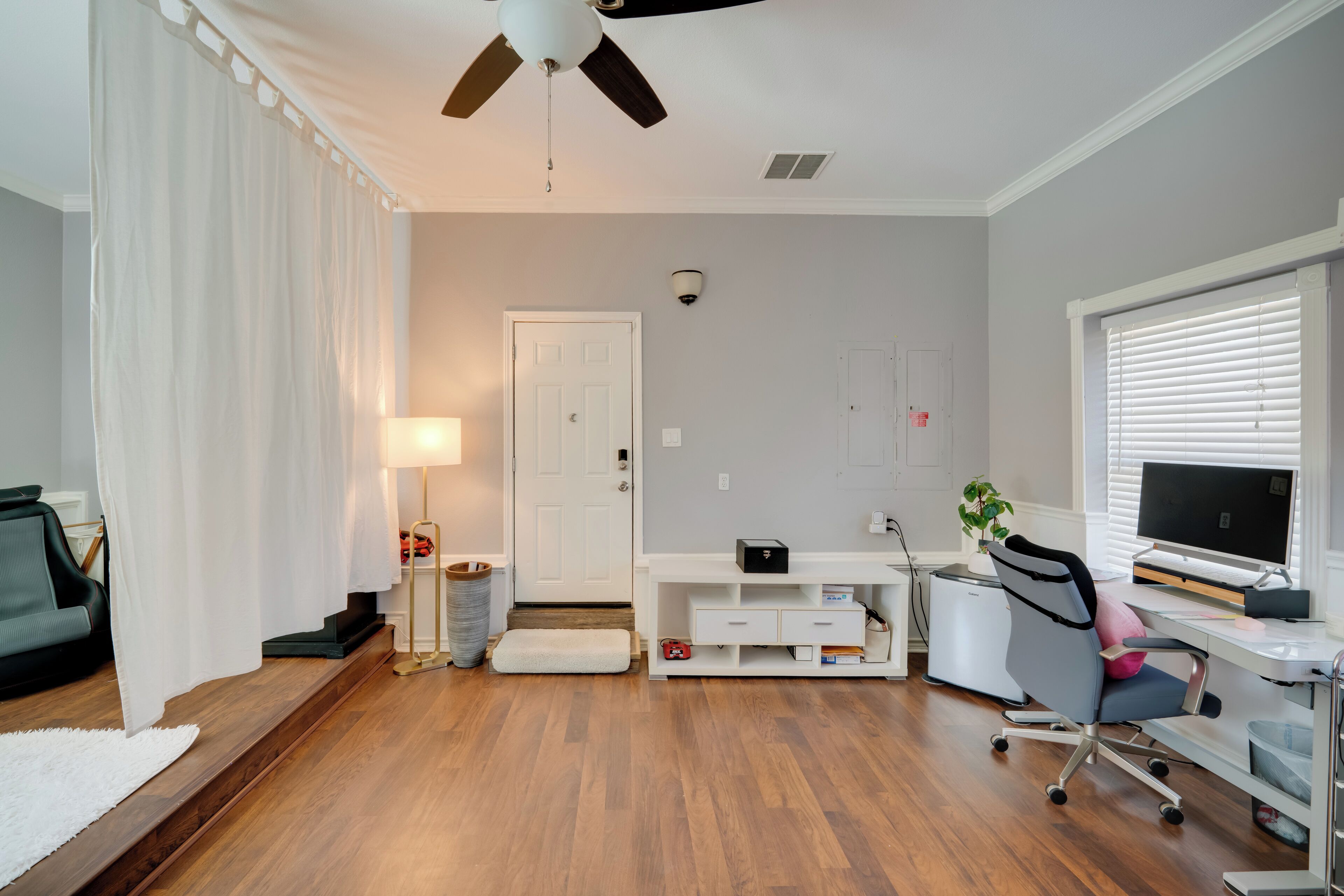 A room with a ceiling fan, a desk with a computer, a chair, a lamp, and a white door.