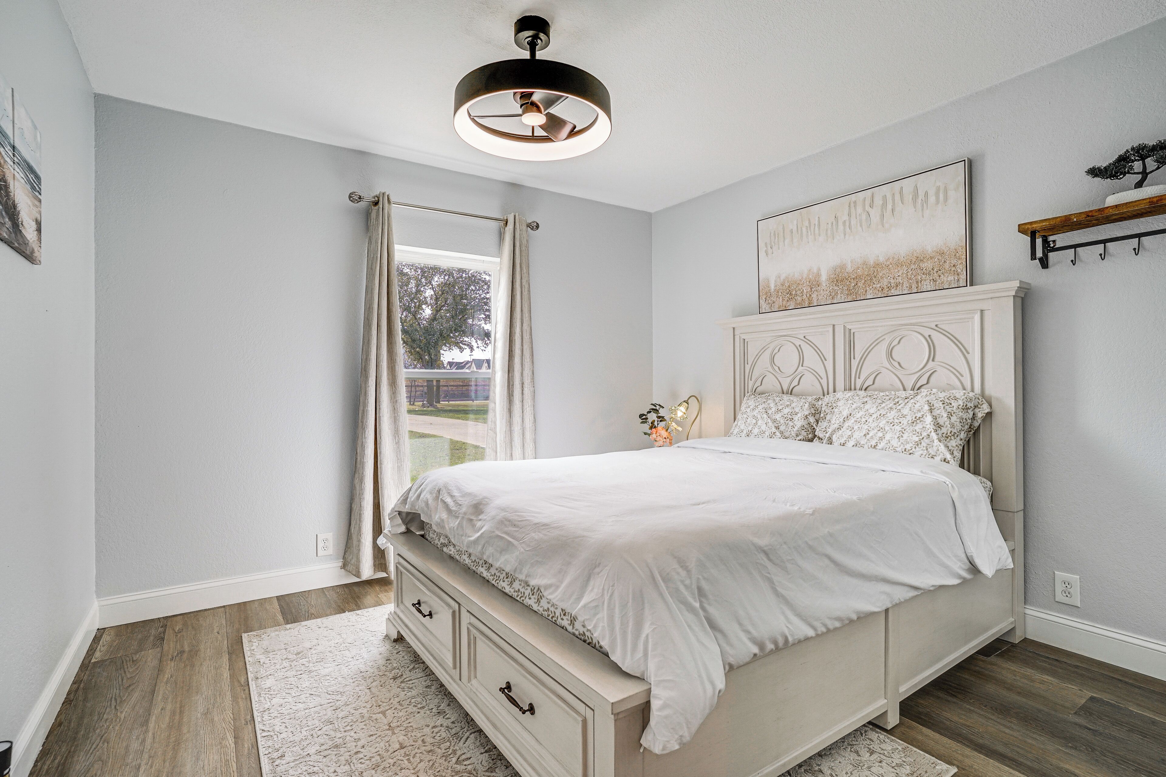 Une chambre à coucher comprenant un lit, un ventilateur de plafond, une fenêtre avec des rideaux, une table de chevet et une étagère fixée au mur.