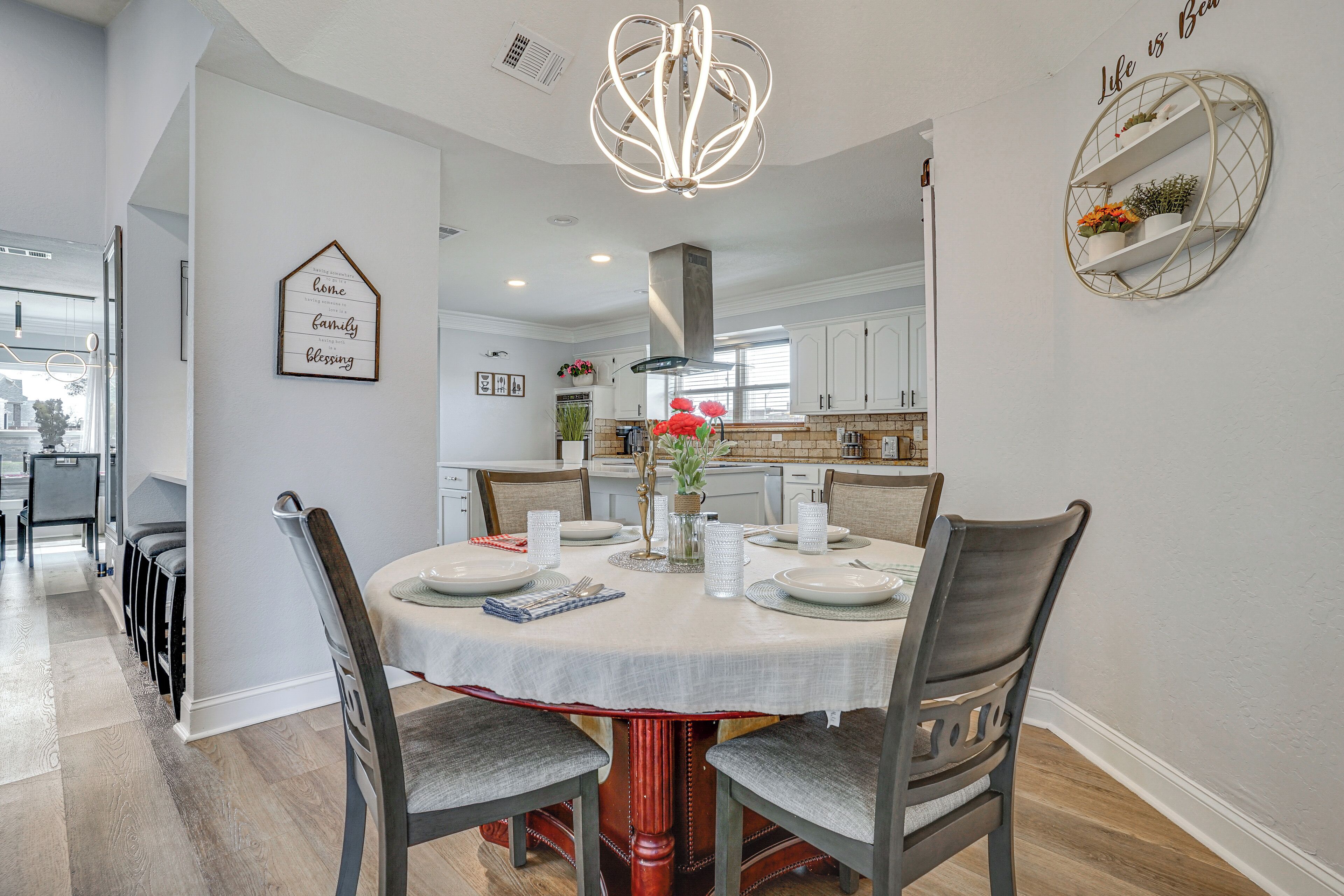 A dining area with a round table set for four, surrounded by chairs. A chandelier hangs above, and there are decorative wall shelves with plants.