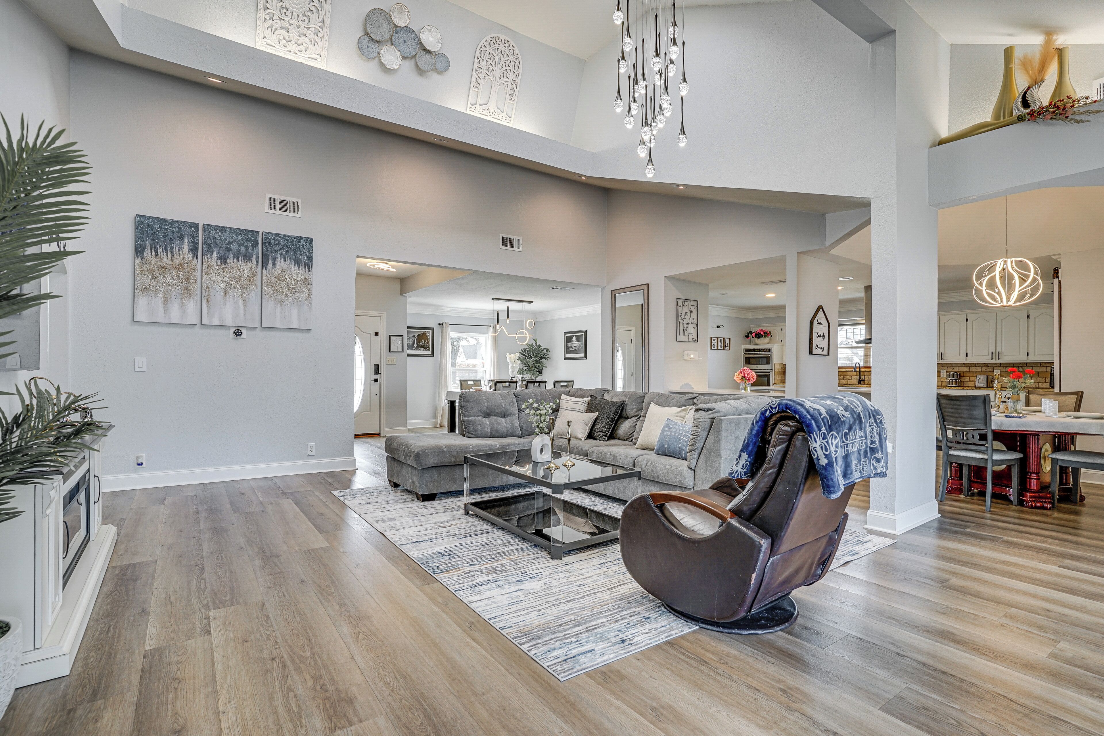 A spacious living room with a gray sofa, a leather chair, and a glass coffee table. There is a dining area with a table and chairs visible in the background.