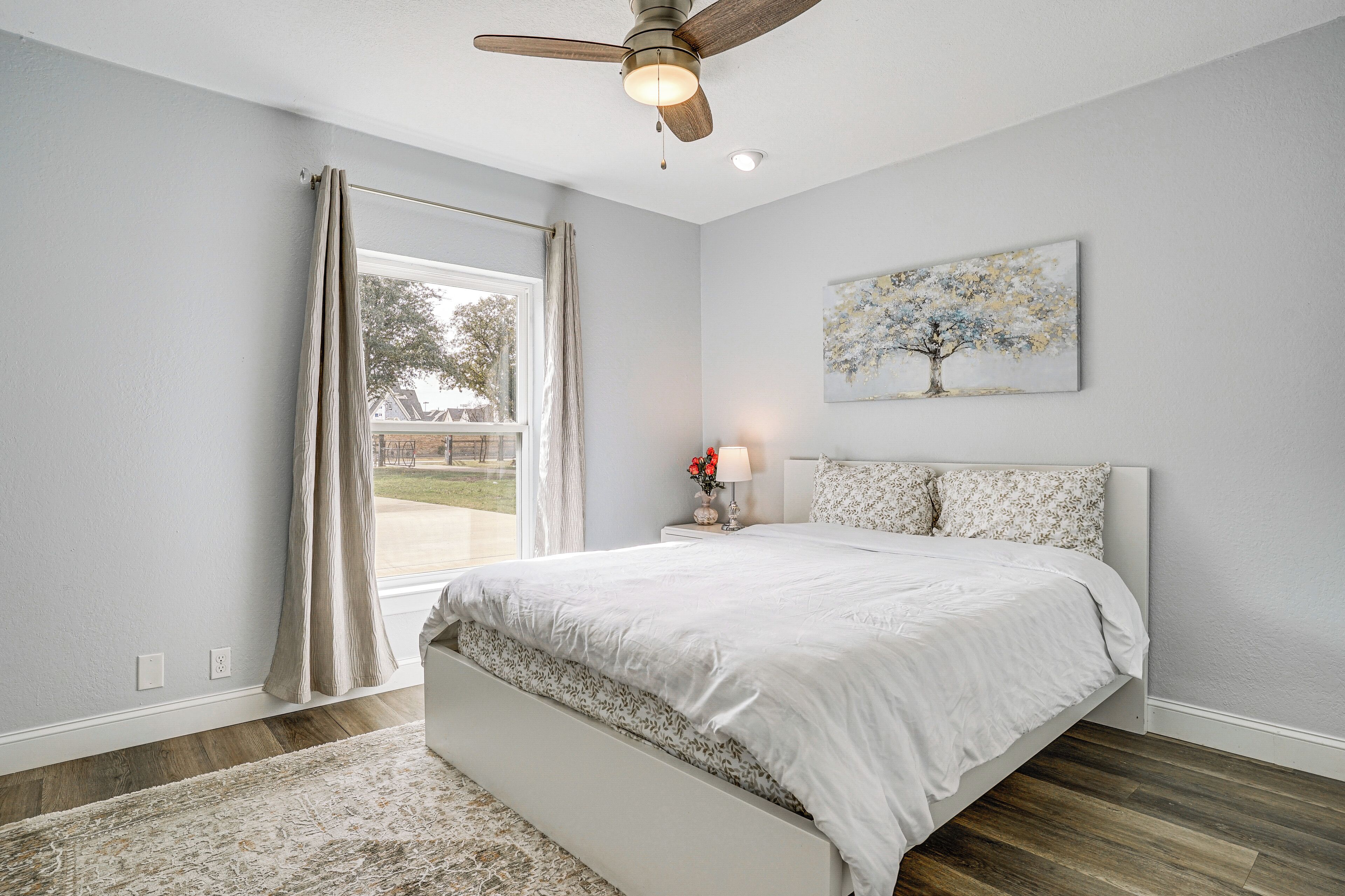 A bedroom with a bed, a ceiling fan, a window with curtains, a nightstand with a lamp, and a painting on the wall.
