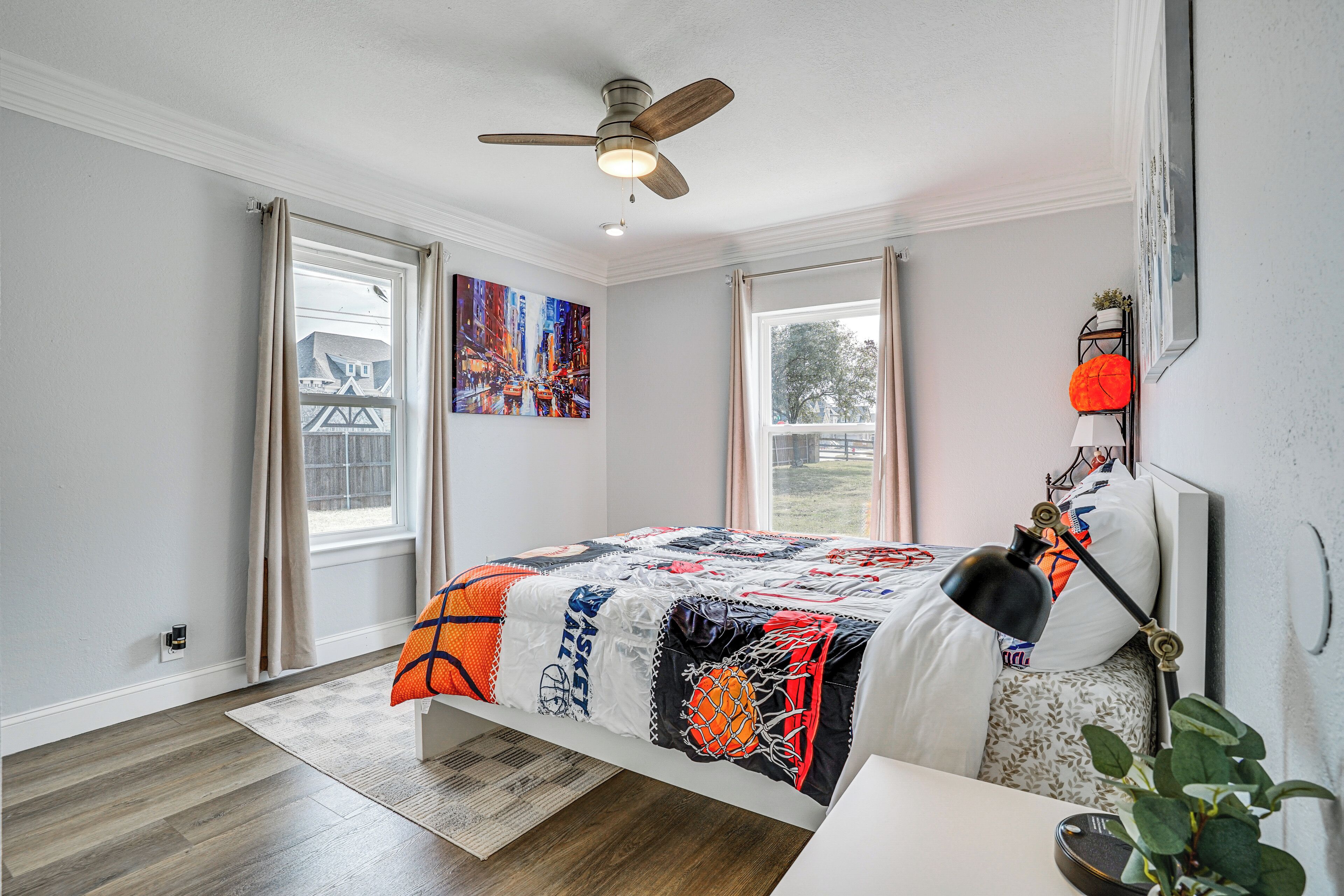 A bedroom with a basketball-themed bedspread, a ceiling fan, and a view of the outdoors through a window.