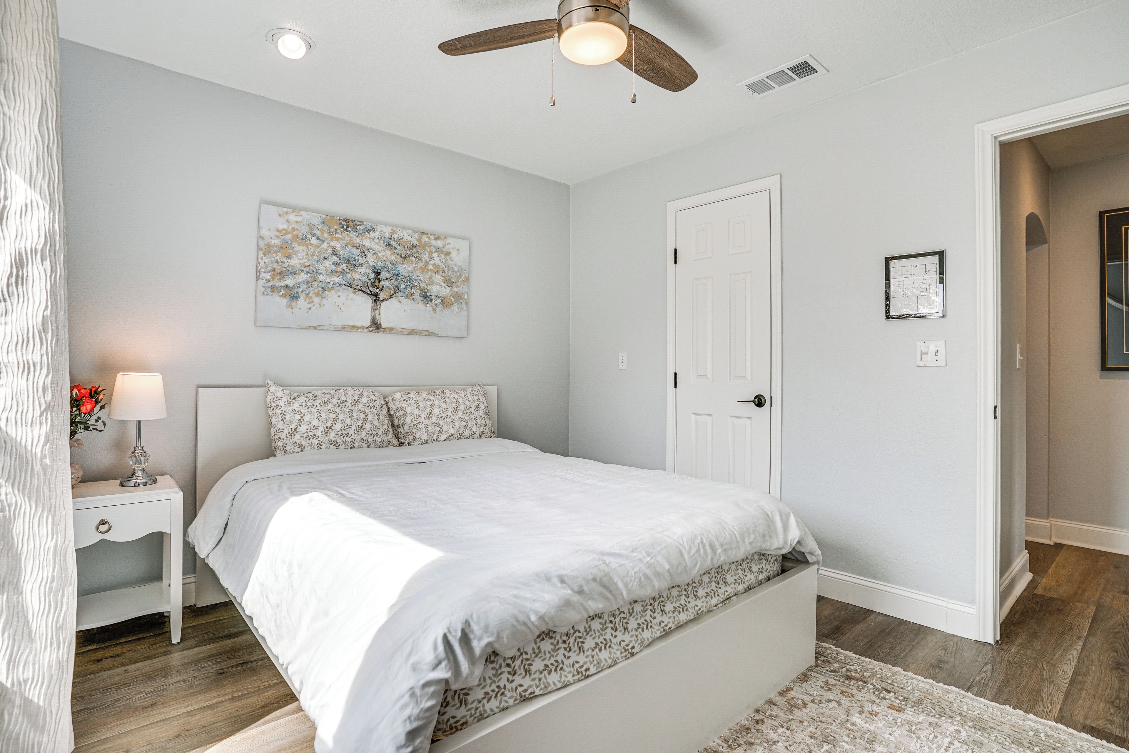 A bedroom with a bed, nightstand, lamp, ceiling fan, and artwork on the wall.
