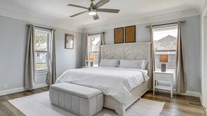 Un dormitorio con cama, mesita de noche, banco, ventilador de techo y ventana con cortinas.
