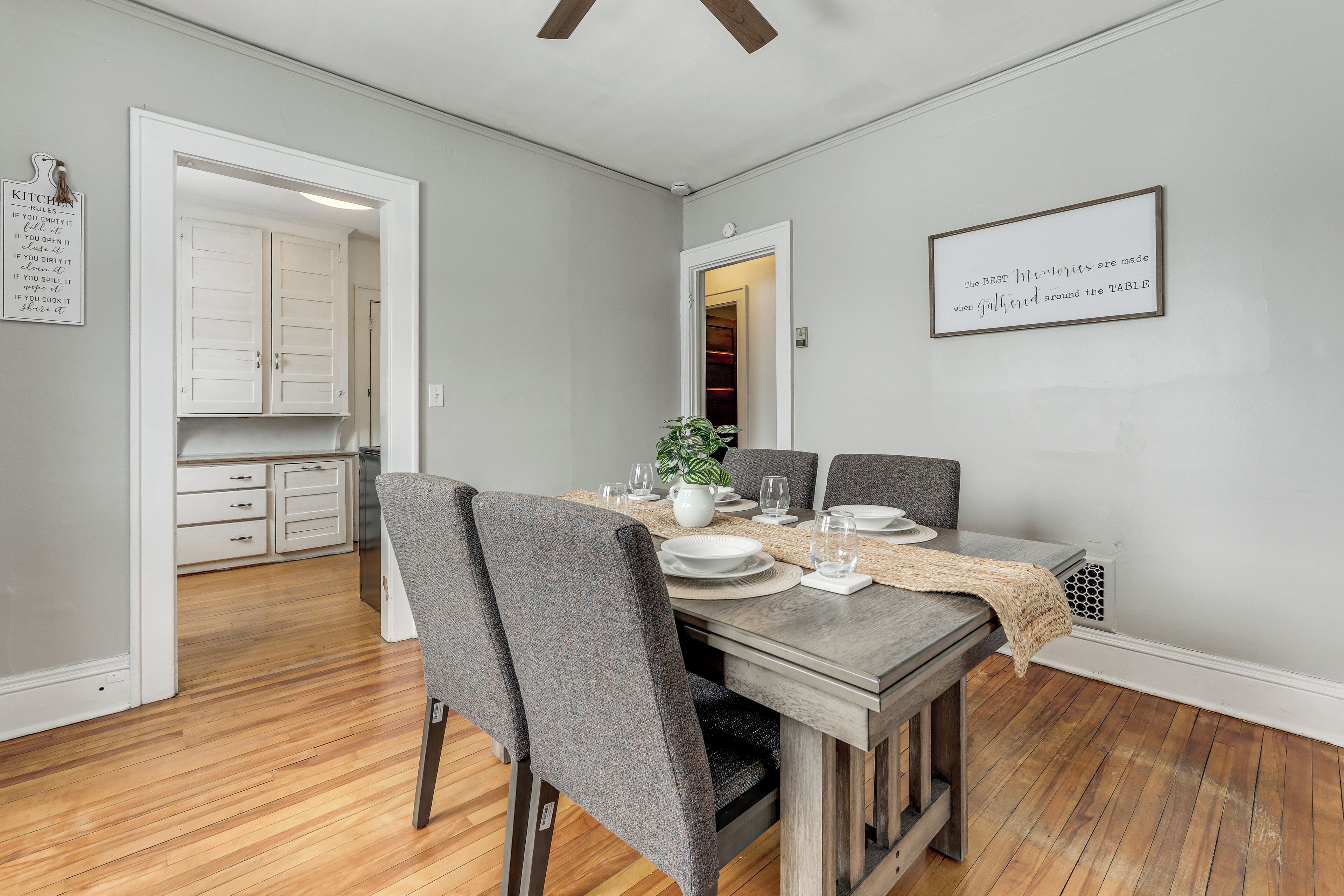 A dining area with a wooden table set for four, grey upholstered chairs, and a kitchen visible through an open doorway.