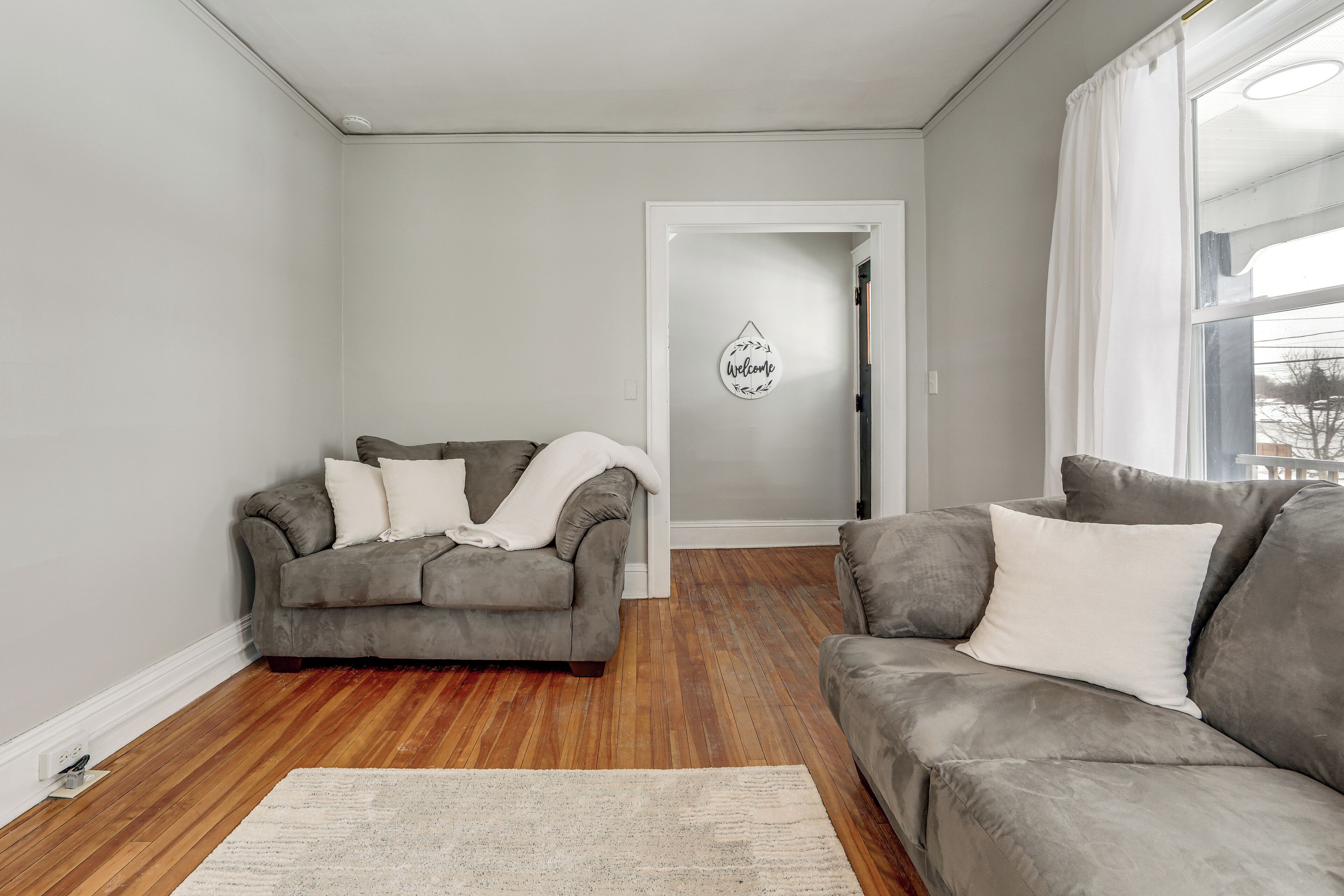 A living room with two grey sofas, a wooden floor, and a 'Welcome' sign on the wall.