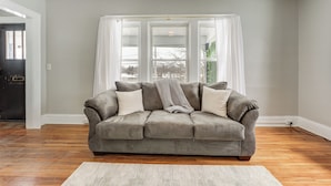 A grey sofa with white cushions in a room with wooden flooring, a white rug, and a window with white curtains.