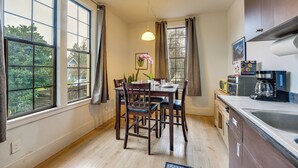 Une cuisine avec des meubles de rangement en bois, une table à manger avec des chaises, un luminaire suspendu et de grandes fenêtres avec des rideaux.