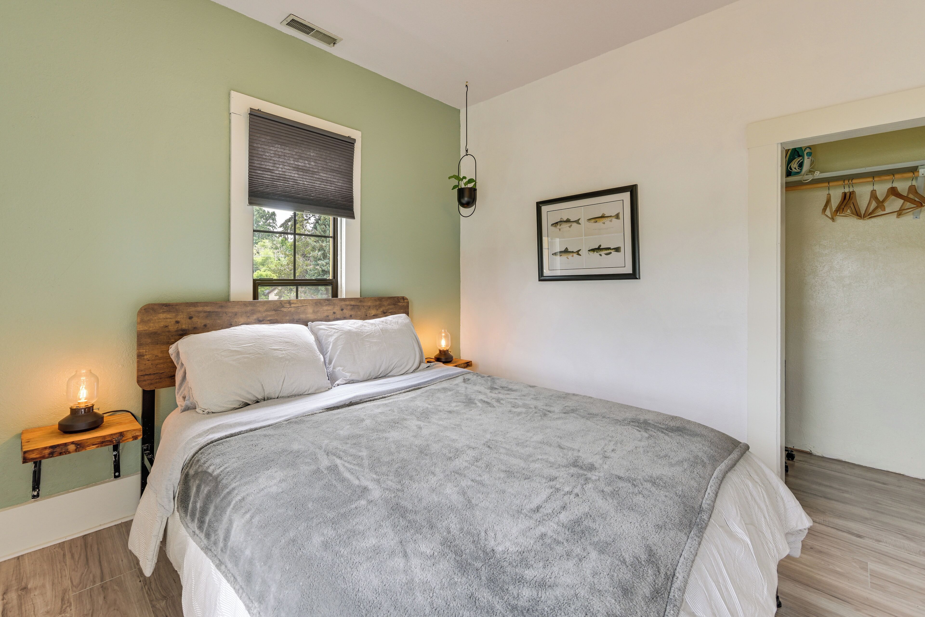 Une chambre à coucher avec un lit, des lampes de chevet, une fenêtre avec des stores, un tableau représentant un poisson encadré et une porte en bois.