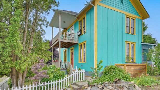 Une maison à deux étages avec un balcon, entourée d’une clôture en bois peinte en blanc, d’arbres et d’un jardin.