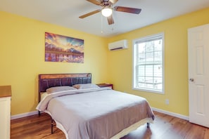 A bedroom with a bed, a ceiling fan, a painting, an air conditioning unit, and a window.