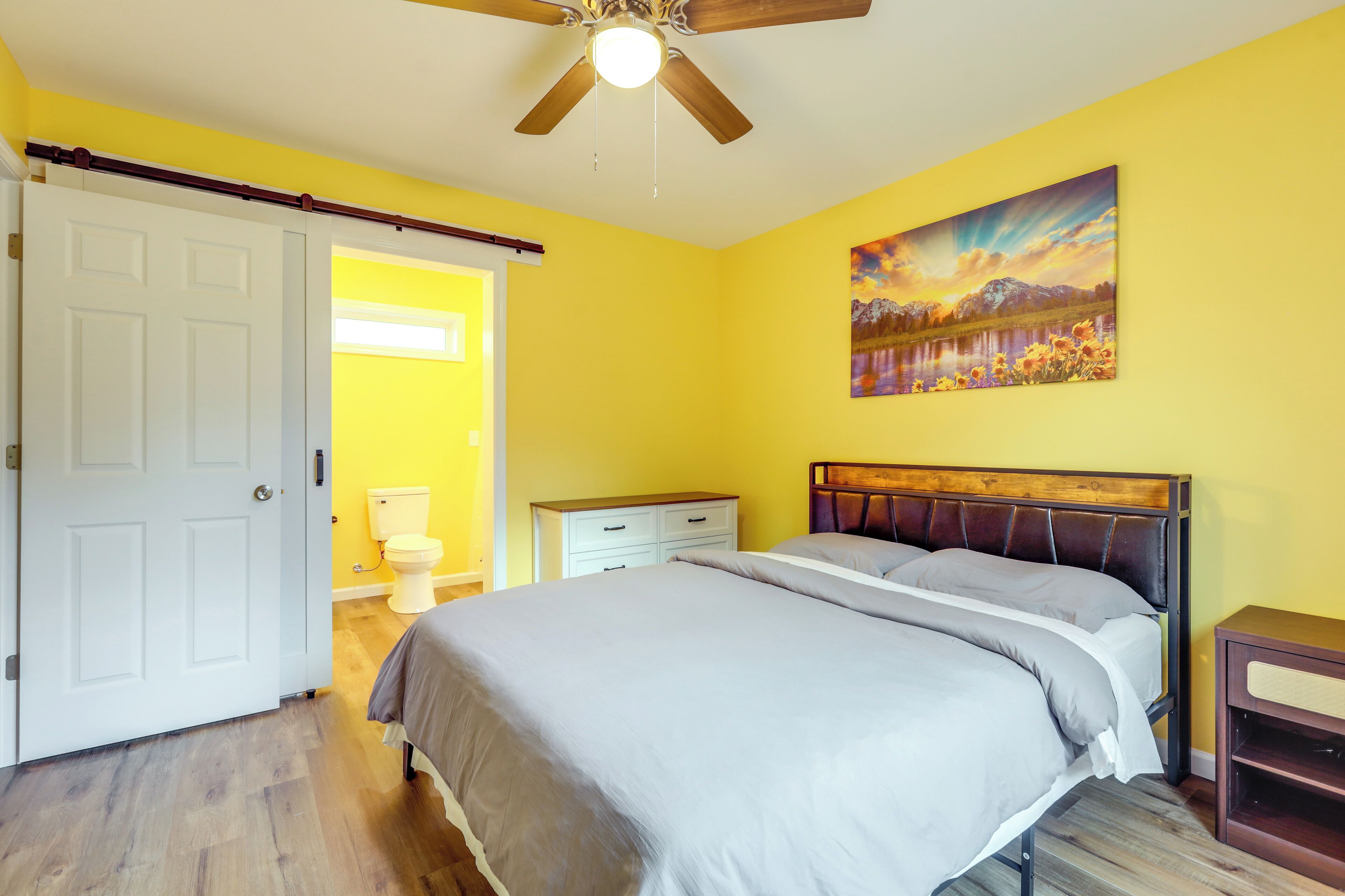 A bedroom with a bed, a ceiling fan, a white door, a white nightstand, and a painting on the wall.