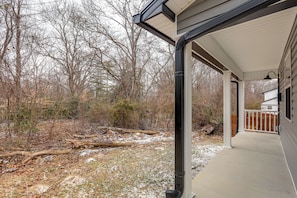 A covered porch with a view of a wooded area and a house in the background.