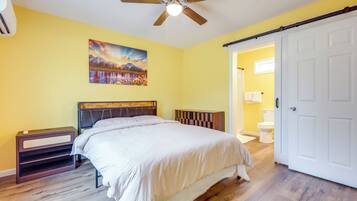 A bedroom with a bed, a ceiling fan, a painting, a nightstand, and a bathroom visible through an open door.