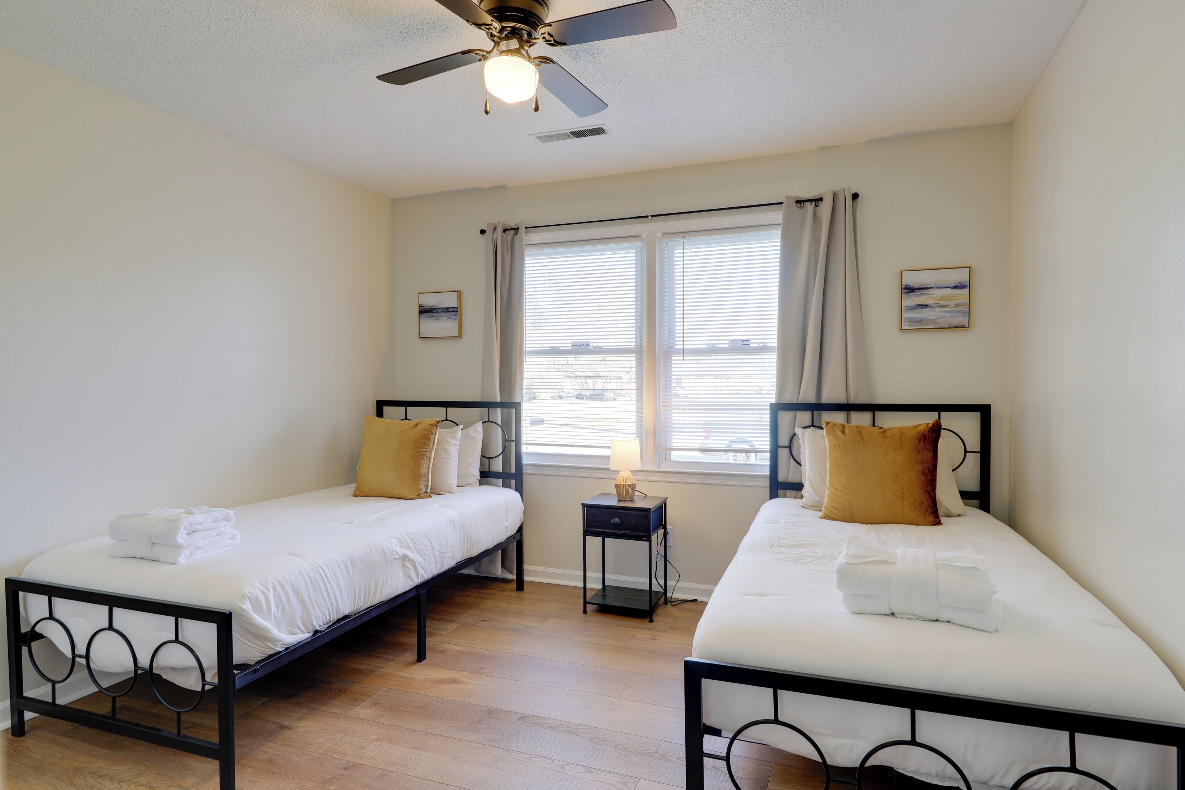 A hotel room with two single beds, a ceiling fan, a nightstand, and a window with curtains.